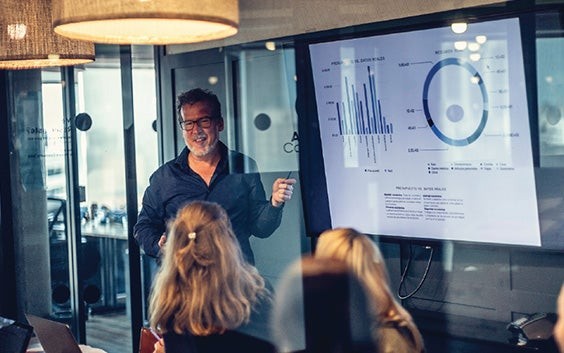 Man talking to a room full of people while pointing to charts on a screen
