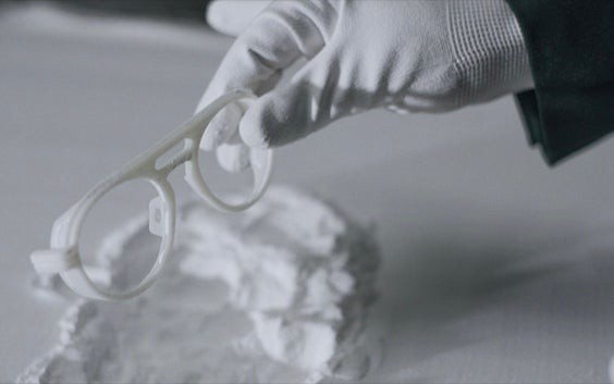 Person wearing gloves and holding 3D-printed eyewear frames above a pile of 3D printing powder