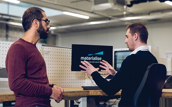 Two men in conversation in front of a computer screen showing the Materialise logo