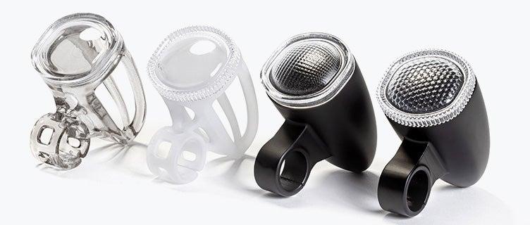 Four 3D-printed bicycle light housings made with VersaClear displayed in a row, showing different shades, from transparent and white prototypes to finished black units with textured reflective lenses.