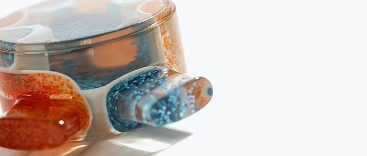 Nahaufnahme eines 3D-gedruckten, durchsichtigen Teils mit orangefarbenen und blauen Partikeln, die auf der Seite verstreut sind